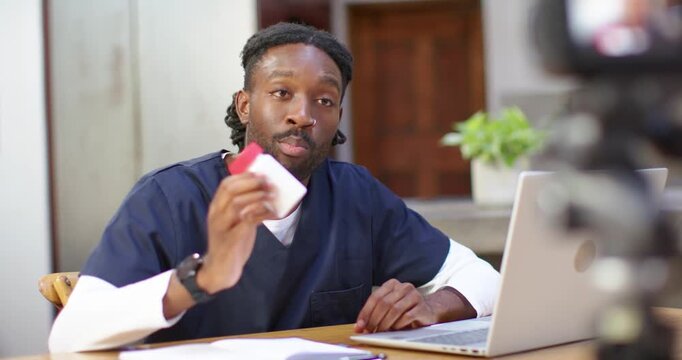African American man sitting in scrubs at desk, camera rolling, holding jar for product demo