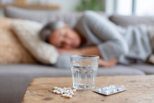 An elderly person rests on a couch, appearing tired or unwell. In focus, a glass of water and scattered pills sit on a wooden table. The scene suggests someone managing illness or medication at home