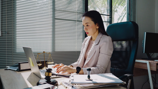 Focused woman works at her legal desk. Hands type on laptop, eyes concentrated. Scales, stamp, and papers hint at legal duties. Office chair, blinds, and monitor complete scene
