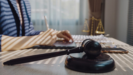 Judge's gavel rests on polished wood. Hands write calmly, focused on legal matters. Sunlight casts sharp stripes across the desk. Scale of justice stands in soft background glow © WARAPHON