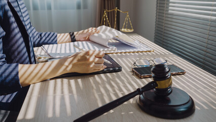 Lawyer sits at desk, hands resting on documents. Sunlight casts sharp stripes across wooden surface. Gavel rests beside smartphone and glasses. Jury scale stands behind, symbolizing justice © WARAPHON