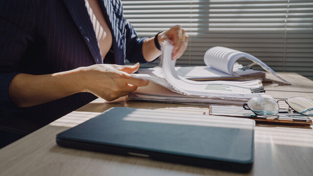 Woman flips through documents at desk. Natural light filters through blinds behind her. Focused, she organizes paperwork with care. Closed laptop rests nearby, ready for use