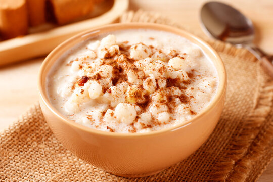 Creamy white corn porridge with cinnamon powder in a bowl on a rustic wooden table. Brazilian dessert with coconut and peanut brittle.