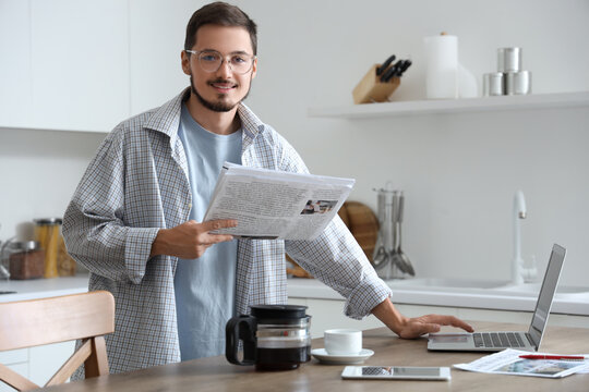 Young man with newspaper using laptop in kitchen