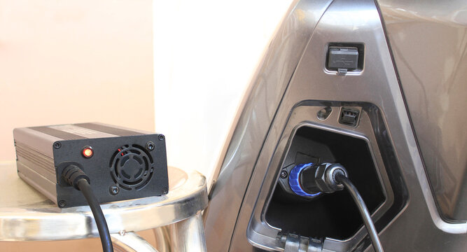 Modern charge EV vehicle electric battery charger plugged into motorcycle port. Clean metallic design highlights advanced technology. Charging port open, cable connected with blue connector.