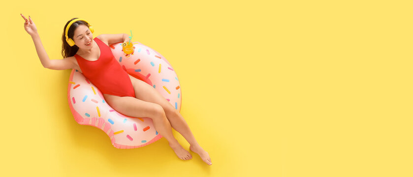 Beautiful Asian woman in swimsuit drinking cocktail and listening to music while relaxing on inflatable ring against yellow background with space for text, top view