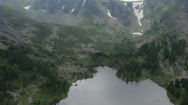 Drone footage of Karakol Lakes in the Altai Mountains, Republic of Altai, Russia, showing calm alpine water, rocky slopes, conifer forest and snow patches
