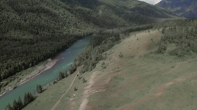 Drone footage of a turquoise river winding through the Ak-Kem valley, with dense taiga forest and green mountain slopes under cloudy sky, Altai Republic, Russia
