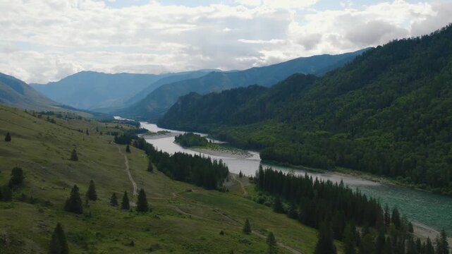 Scenic aerial footage of a winding river flowing through Ak-Kem valley, with forested hills and mountain ridges under dramatic clouds, Altai Republic, Russia
