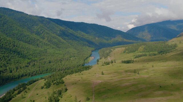 Drone footage of the Ak-Kem valley with a turquoise river winding between forested mountains and open meadows, Altai Republic, Russia, under cloudy summer sky