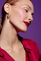 Fototapeta premium Close-up of a woman with glowing skin and red lipstick wearing gold earrings and a red blouse against a purple background. Concept of beauty, makeup, and fashion.