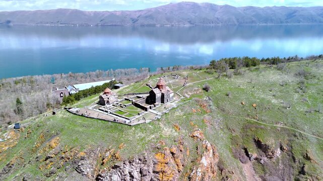 Aerial view of Sevanavank monastery complex on a green peninsula with tourists walking around the stone ruins overlooking the blue waters of Lake Sevan, Sevan, Gegharkunik Province, Armenia.