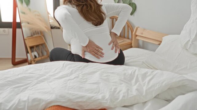 Woman sitting on bed with hands on lower back, wincing and adjusting posture beside mirror and rattan chair in a building bedroom; discomfort recovery.