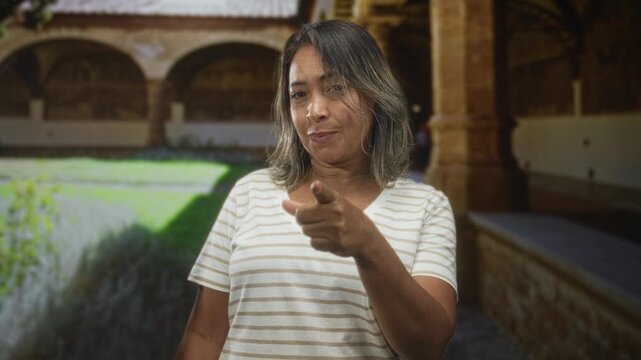 Hispanic woman points finger at camera in historic cloister building, striped v neck shirt, middle age, direct gaze, pointing gesture; confidence empowerment.