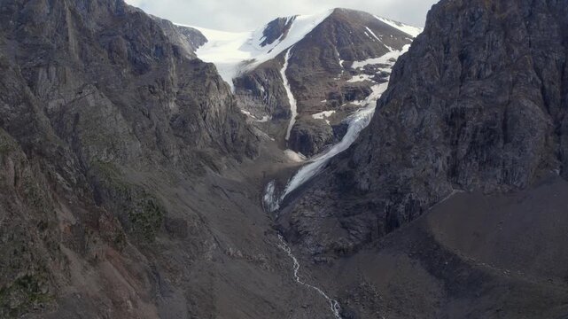 Drone footage of Aktru Valley with glacier tongue, steep rocky cliffs and snowfields in the Altai Mountains, Siberia, Russia, under cloudy summer sky