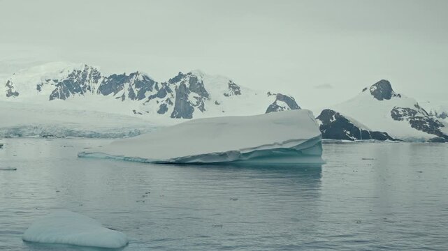Large flat-topped tabular iceberg with blue glacial layers drifts on calm polar ocean surrounded by brash ice and dramatic snow-covered Antarctic mountains