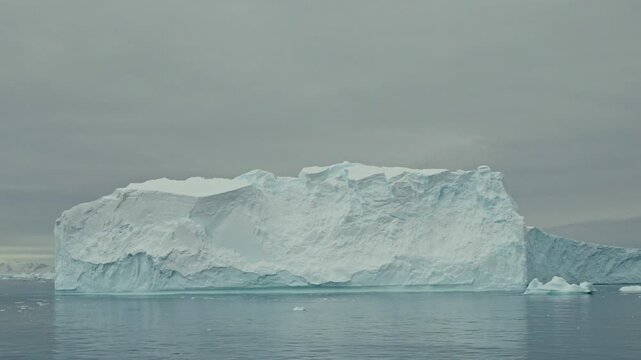 Massive flat-topped tabular iceberg with dramatic eroded cliff walls and blue glacial texture floats on calm dark polar ocean under heavy grey sky