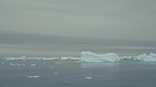 Wide minimalist shot of scattered icebergs and brash ice drifting silently across vast open polar ocean under heavy uniform grey overcast Antarctic sky