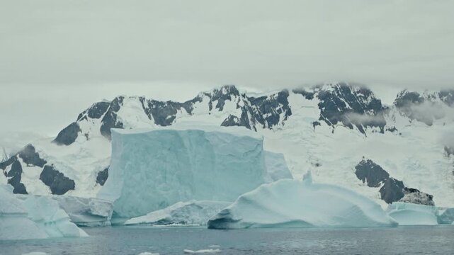 Slow pan past dominant tabular iceberg and layered ice formations against dramatic snow-patched rocky Antarctic mountain coastline