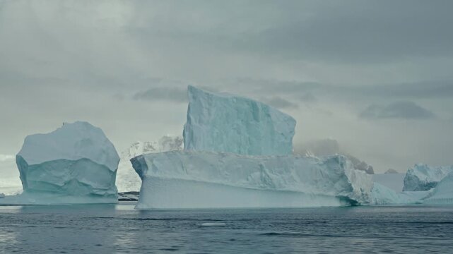 Orbiting massive blue-tinted tabular icebergs drifting in dark Antarctic ocean with misty snow mountains under dramatic overcast sky