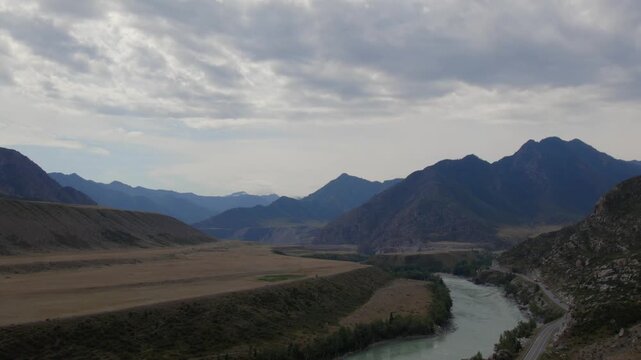 Drone footage of Chuysky Trakt highway winding through a wide valley beside a turquoise mountain river and rugged peaks, Altai Republic, Russia under cloudy sky