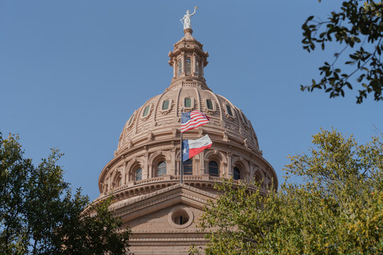Front facade of the Texas State Capitol building in Austin, with USA and Texas flags, surrounding trees, and clear blue sky.