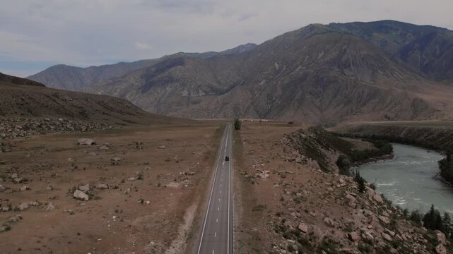 Drone footage of the Chuysky Trakt highway cutting through a wide mountain valley beside a turquoise river in Altai Republic, Russia, under cloudy sky