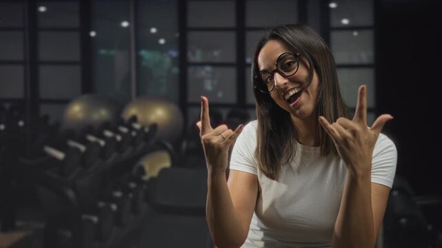 Woman in studio shows rock horns with hands and glasses, smiling in a white shirt with dumbbells visible behind her; playful confidence.
