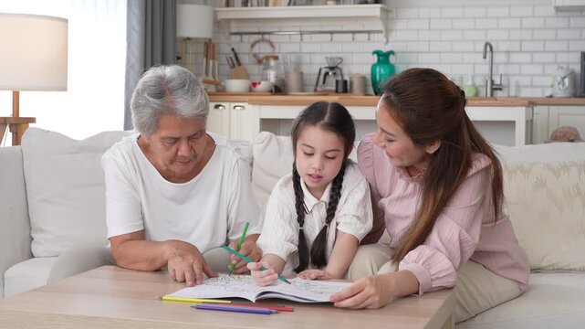 Happy Asian three generation family drawing together in living room, Smiling grandmother, mother and little daughter coloring book on table, Warm home education, bonding and family leisure concept