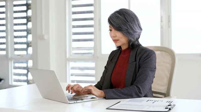 Mature senior asian professional business woman working on laptop, looking at screen searching information at office.