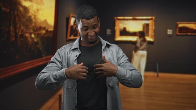 Young black man with palms raised in stop gesture beside framed paintings in museum gallery; playful caution.