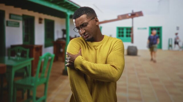 Young man clutching shoulder while wincing in a sunlit building courtyard wearing yellow sweater and glasses; discomfort concern.