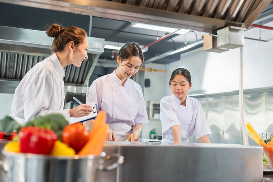 Professional female chef teaching young students in industrial kitchen classroom, Culinary instructor supervising Asian students during cooking class in restaurant kitchen
