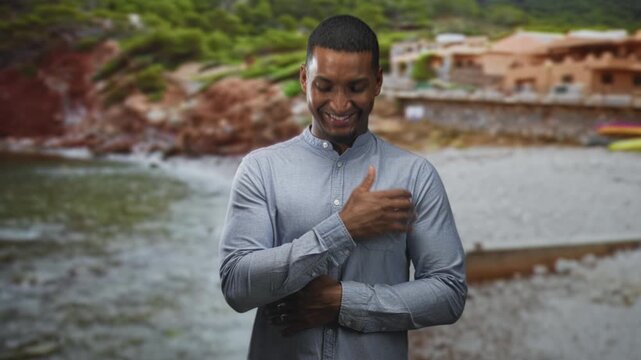 Black man with hand on chest smiling, other arm folded across torso, standing in front of a rustic building on a pebbled beach by the sea; gratitude serenity.