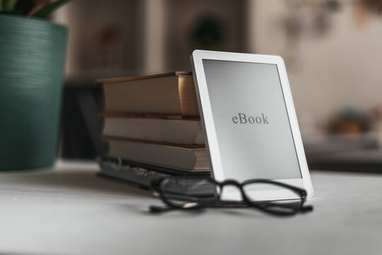 Ebook reader and glasses on desk with books and plant in soft home office setting. Cozy workspace, remote work lifestyle and calm productivity concept with minimalistic interior design.