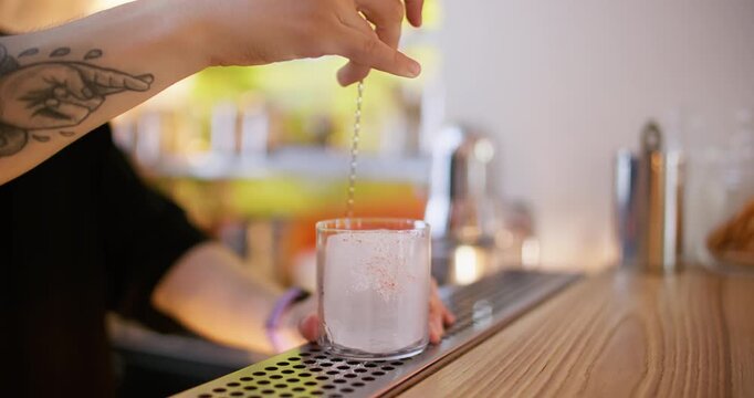 Drink making. Cold cocktail. Bartender hand stirring ice cubes in glass with bar spoon preparing chilling ingredient on pub counter.