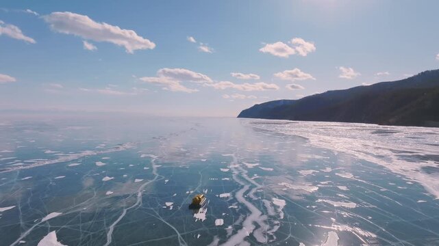 Aerial view of a hovercraft traveling across cracked blue ice on Lake Baikal in Siberia, Russia, with snowy shoreline and mountains on a clear winter day