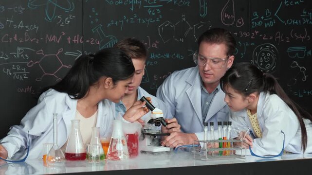 Multicultural student looking under microscope while cute girl inspect sample. Teacher looking after high school children doing experiment at blackboard with science theory in STEM class. Edification