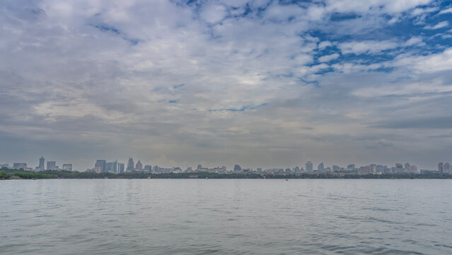 A large calm urban lake. Ripples on the water. On the far shore there is green vegetation, city buildings, skyscrapers. Blue sky, clouds. China. Hangzhou. West Lake Xi Hu