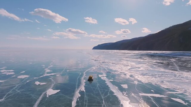 Aerial winter footage of a hovercraft traveling across the frozen ice of Lake Baikal with natural cracks and snow patterns, mountains on the shore, Russia