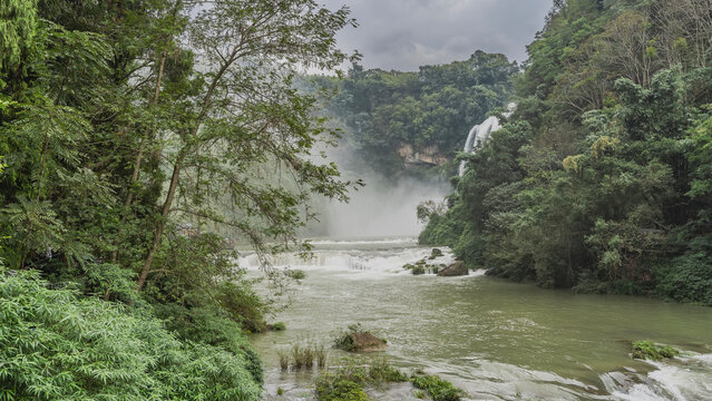 The river flows between the mountain slopes. Foam on river rapids. Streams of the waterfall flow down from the cliff. Splashes, fog. Lush green vegetation. China. Huangguoshu Waterfall. Guizhou