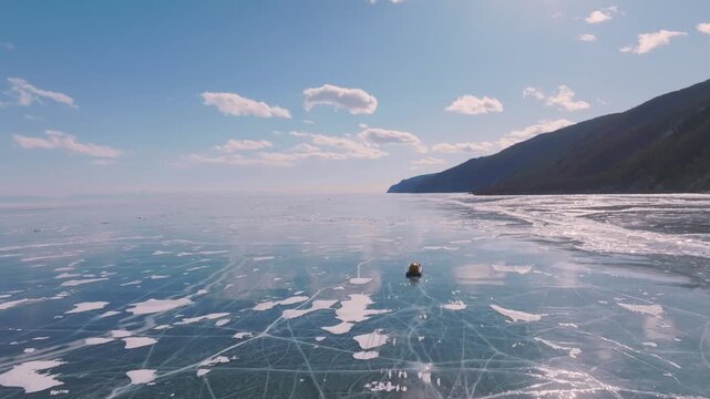 Aerial footage of a hovercraft traveling across the clear cracked ice of Lake Baikal in winter, with mountain shoreline and blue sky, Russia