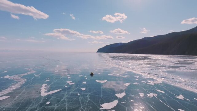 Wide winter landscape footage of a hovercraft traveling across clear cracked ice on Lake Baikal, Russia, with snowy shoreline and mountains under blue sky
