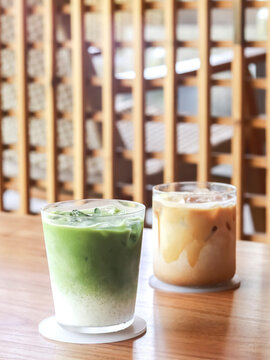 Two refreshing iced beverages on a wooden table in a cozy cafe setting: a layered iced matcha latte with creamy milk and a smooth iced coffee latte.