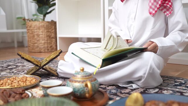 Arab muslim boy hands holding and reading the Holy Quran green book, Wearing traditional white thobe and red keffiyeh, Concept of Islamic religion, faith, Ramadan culture and education