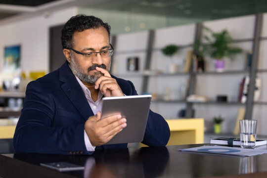Thoughtful businessman sits in coworking office, reviewing report on digital tablet, analyze information, considering next steps, making business decision, planning strategy, thinks on financial data