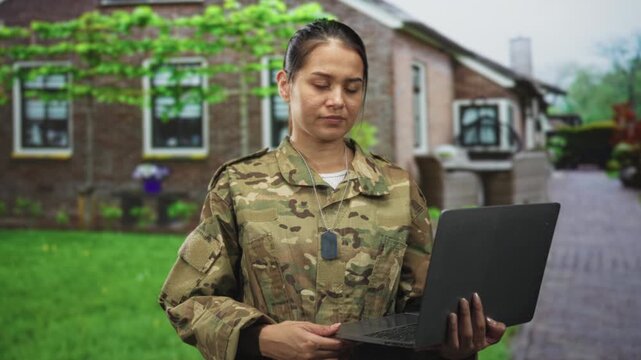 Woman soldier in camouflage holds a laptop with dog tags visible while standing on a brick path by a building; duty resilience.