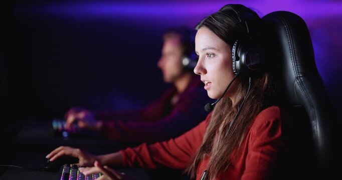 Young adult female gamer wearing headset, sitting at computer, playing streaming video game, immersed in competitive e-sports match in modern cybercafe. Online gaming activity, hobby, entertainment