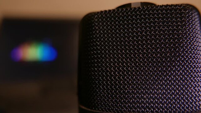 Podcast studio: slow zoom in and close-up shot revealing a professional microphone. Colorful sound levels moving on a laptop screen in the background. POV of a podcast microphone. Podcast gear. A mic.