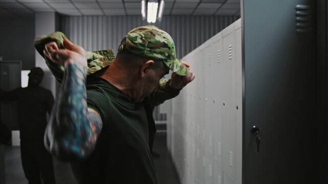 Male Soldier With Tattooed Arms Puts on Camouflage OCP Uniform Jacket and Cap by Locker, Getting Ready For Tactical Mission While Colleagues Interact in Background. Concept Military Base Preparation.